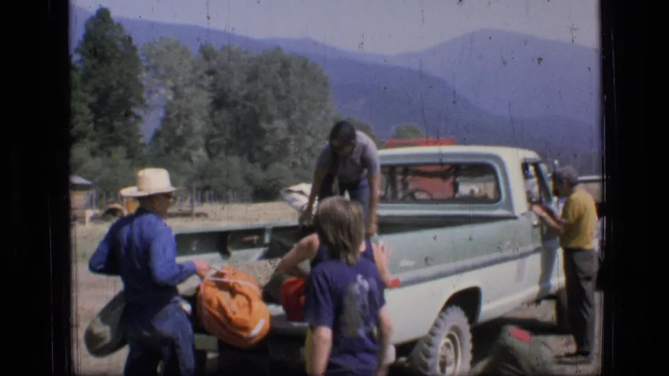 A person carries an orange bag from a light green pickup as others stand nearby in 1973 Montana wilderness footage.