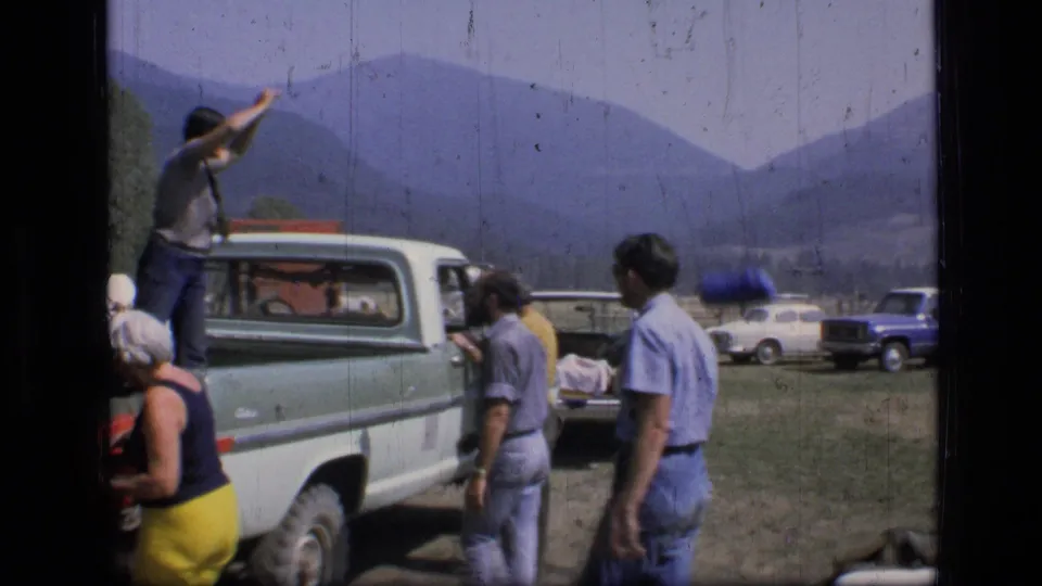 Home-movie frame from 1973 Montana showing a man in a pickup bed near campers, parked vehicles, meadow, and distant mountains.