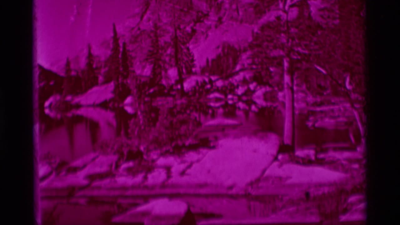 1975 8mm view of Minaret Wilderness showing a magenta-tinted forested lake edge with rocks and still water reflections.