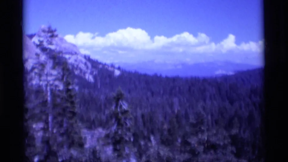Forested Sierra Nevada mountain valley in the 1975 Minaret Wilderness home-movie footage, with ridges, trees, blue sky, and clouds.