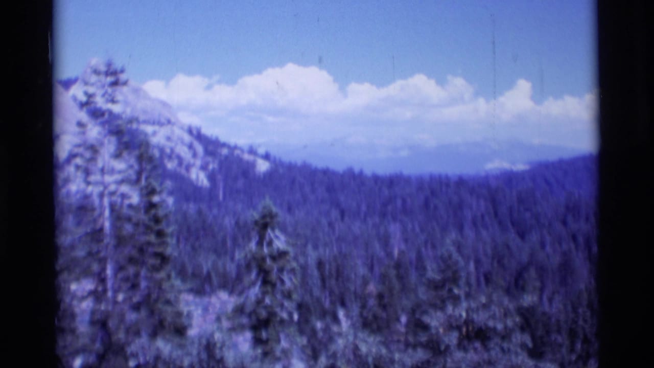 1975 8mm view of the Minaret Wilderness area with evergreen forest, distant ridges, pale clouds, and dark film-frame edges.