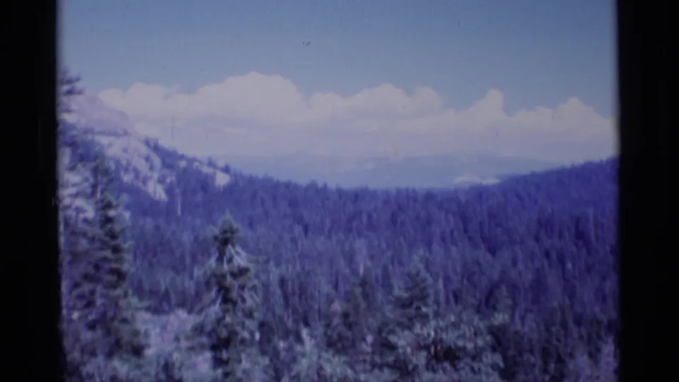 Archival 8mm frame of California wilderness terrain, with tree-covered ridges, distant mountains, blue sky, and soft film grain.