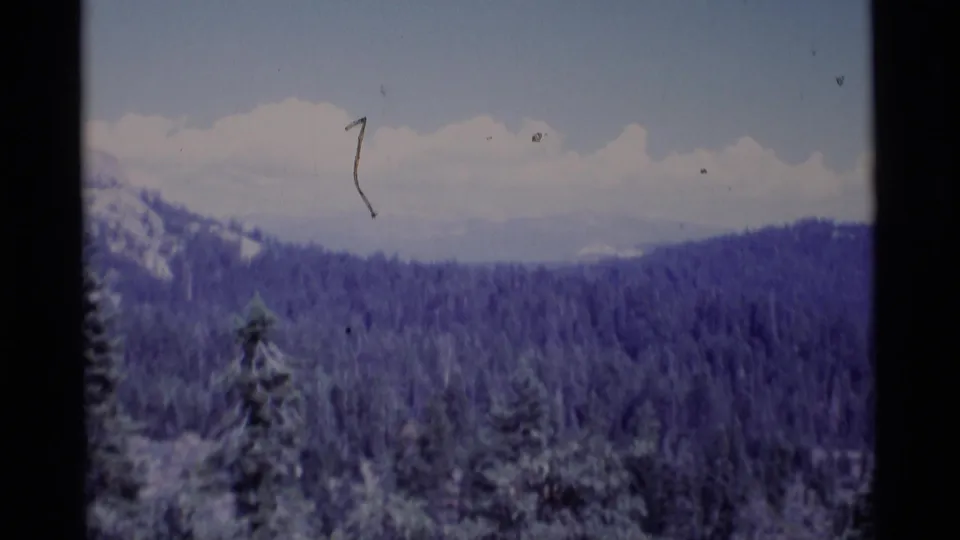 1975 home-movie frame showing Minaret Wilderness forest and mountain ridges, with visible scratches, grain, and a purple-blue cast.