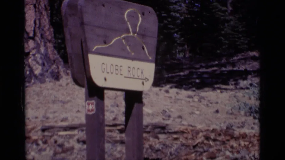A wooden Globe Rock trail sign with an arrow stands beside pine trees and rocky ground in the 1975 Minaret Wilderness footage.