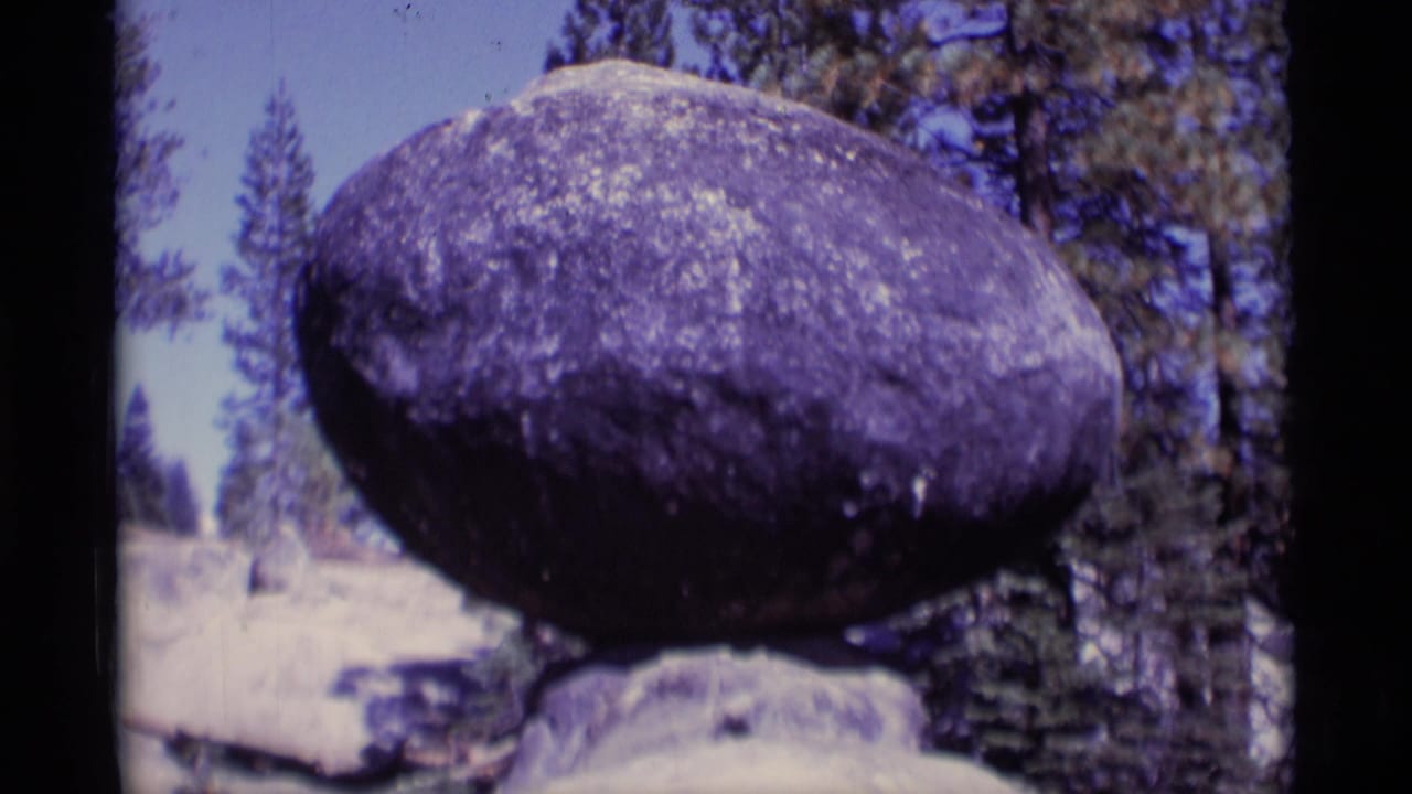 Large rounded boulder balanced on a smaller rock among pine trees.