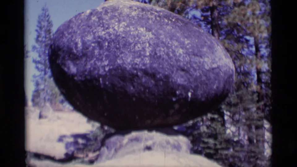 The balanced rock formation remains centered, with pine trunks, sky, and 8mm edge borders visible around the frame.