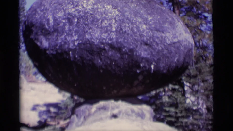 Closer 8mm view of the purple-shifted balanced boulder, with pine trees and pale ground partly visible behind it.