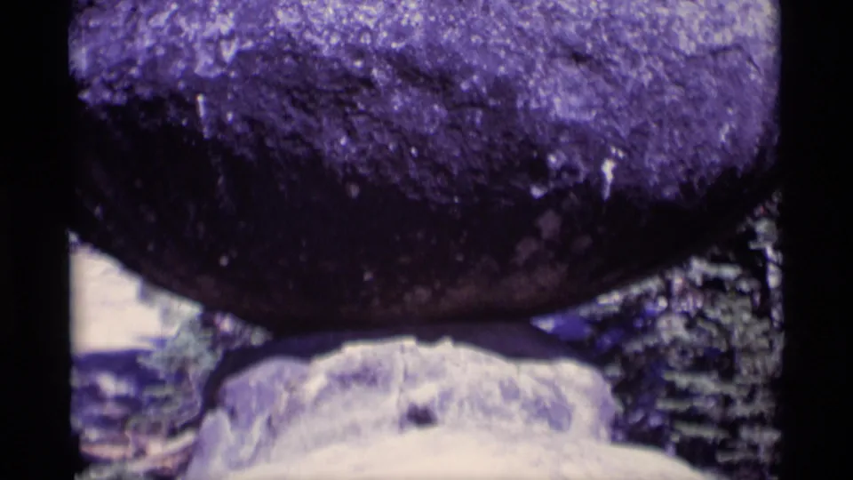 Close 1975 8mm frame showing the balanced boulder dominating the view, with its smaller base rock and forest background still visible.