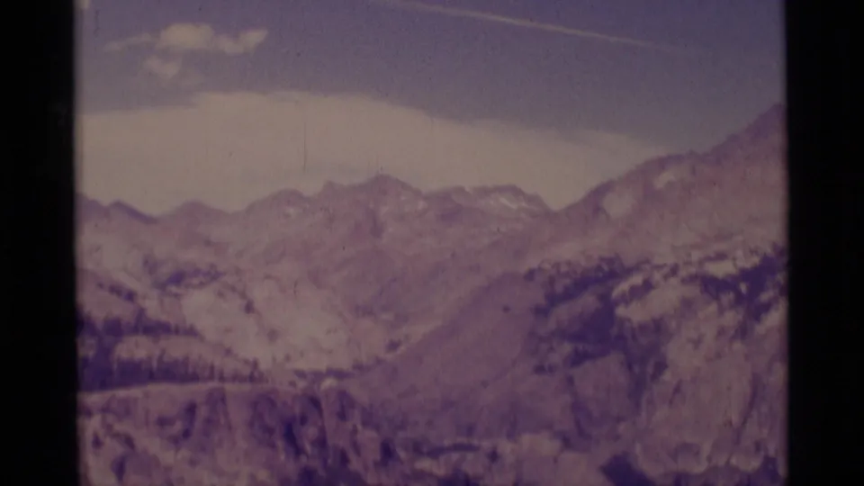 Wide 1975 8mm view across Minaret Wilderness mountain peaks, with snow patches and a faint contrail in the pale sky.