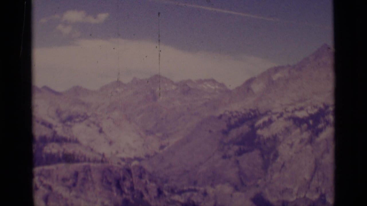 Distant mountain ridges with snow patches under a pale sky on aged 8mm film.