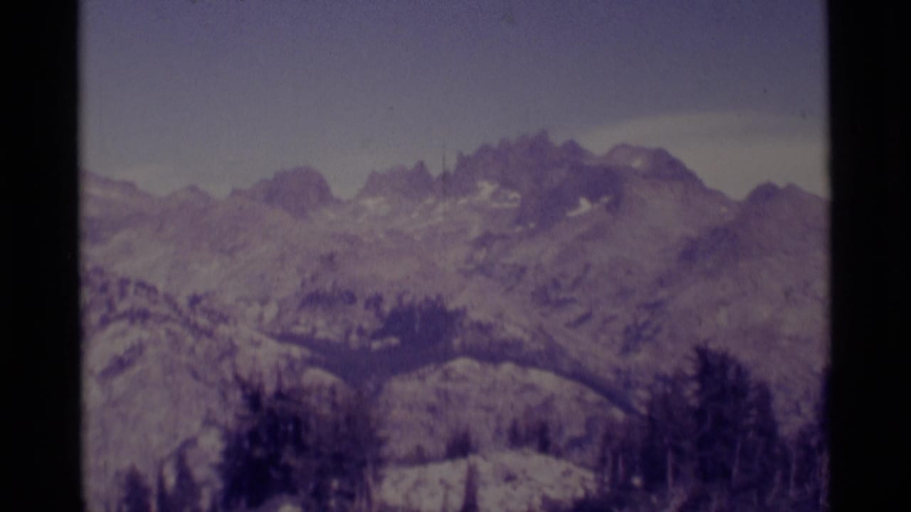 1975 8mm mountain panorama in the Minaret Wilderness area, with muted color, rocky ridges, snow traces, and foreground trees.