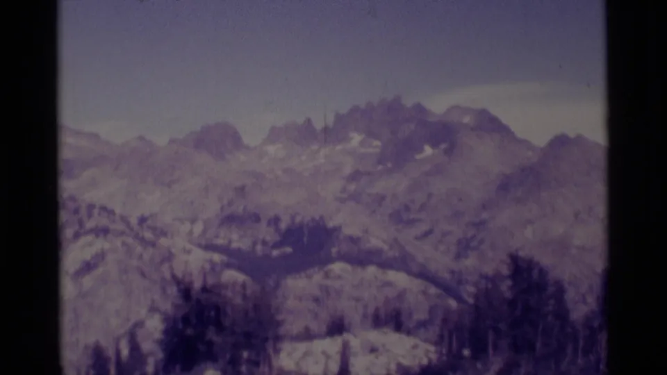 1975 8mm mountain panorama in the Minaret Wilderness area, with muted color, rocky ridges, snow traces, and foreground trees.
