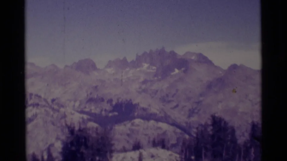 Muted 1975 8mm view of California alpine ridges, scattered snow, gray sky, and dark trees at the base of the slopes.