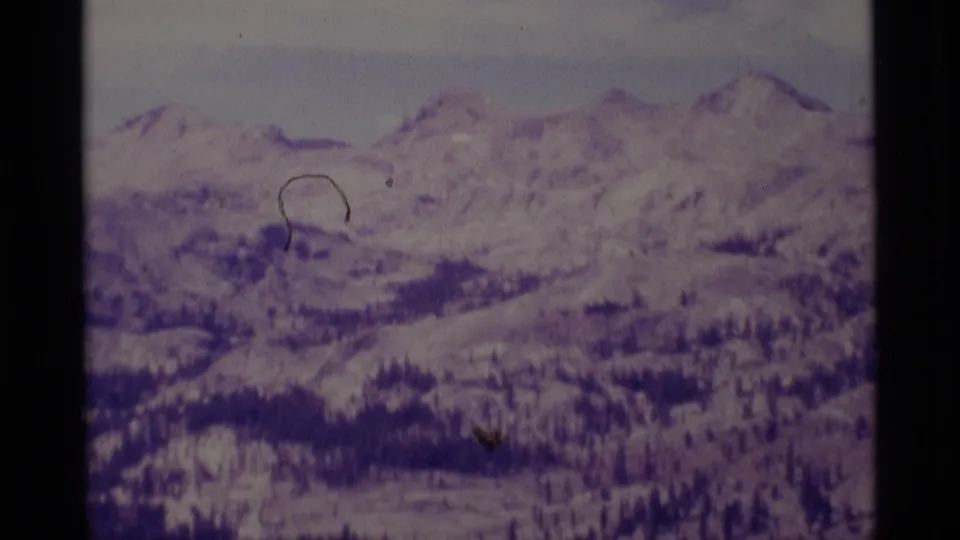 1975 8mm frame of Minaret Wilderness mountain terrain, with snowy peaks, evergreen patches, and visible scan scratches.