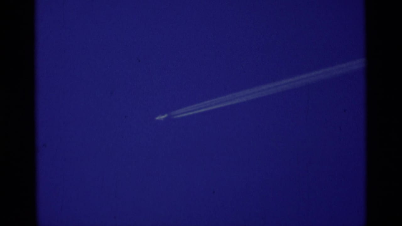 A small aircraft contrail cuts across a saturated blue sky in 1975 8mm Minaret Wilderness footage.