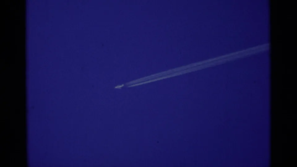 A small aircraft contrail cuts across a saturated blue sky in 1975 8mm Minaret Wilderness footage.