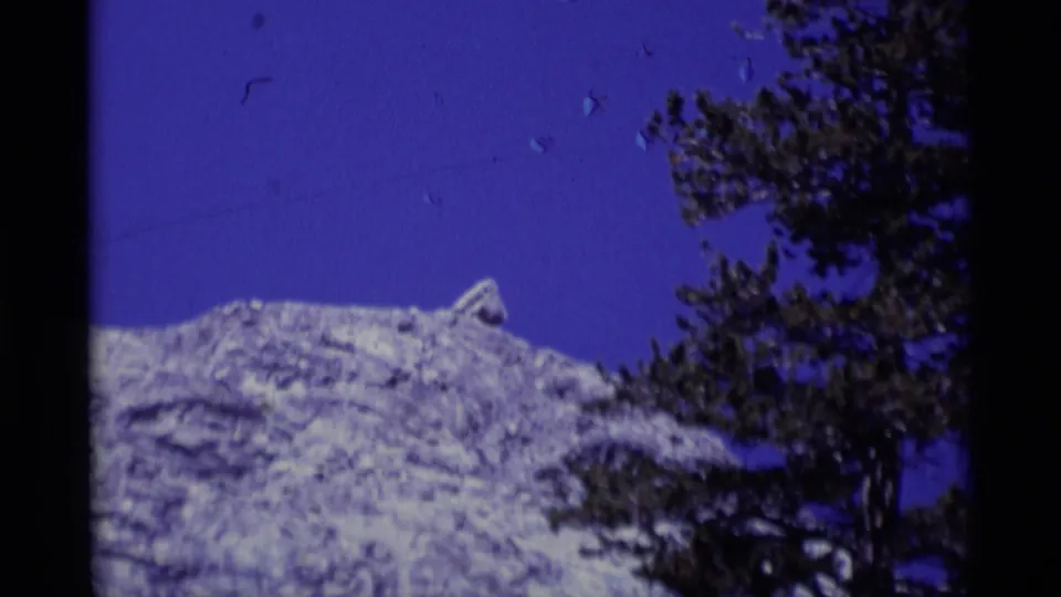 1975 8mm California wilderness frame with pale rock, saturated blue sky, and dark pine trees along the right edge.