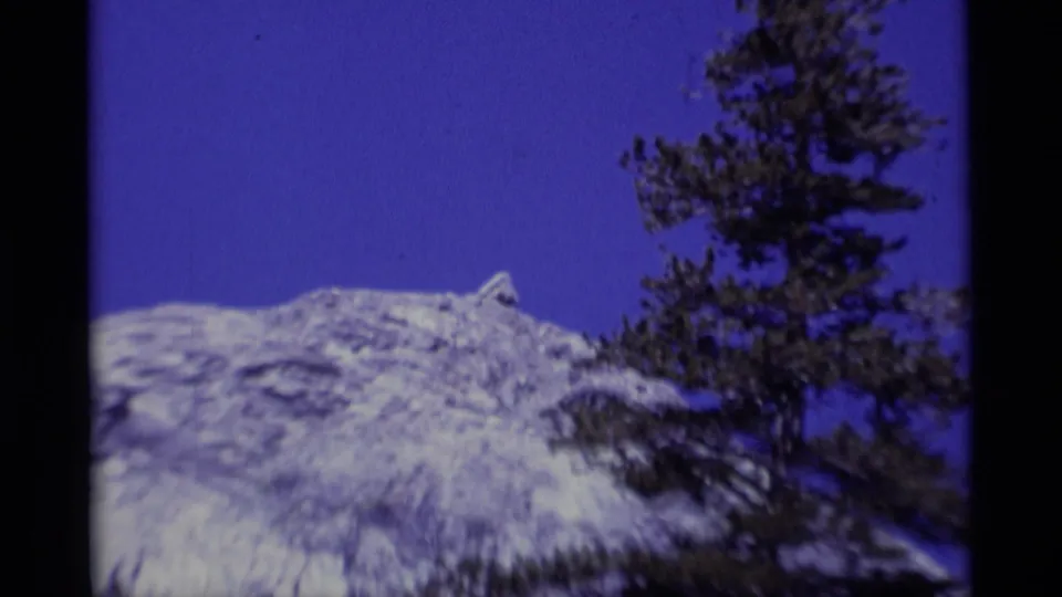 1975 8mm Minaret Wilderness frame showing a pale rocky ridge and a foreground pine against a saturated blue sky.