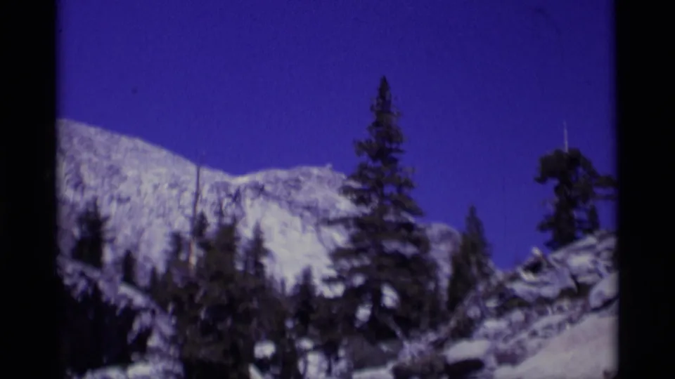 1975 8mm view of Minaret Wilderness with a snowy mountainside, scattered evergreens, and dark film-frame edges.