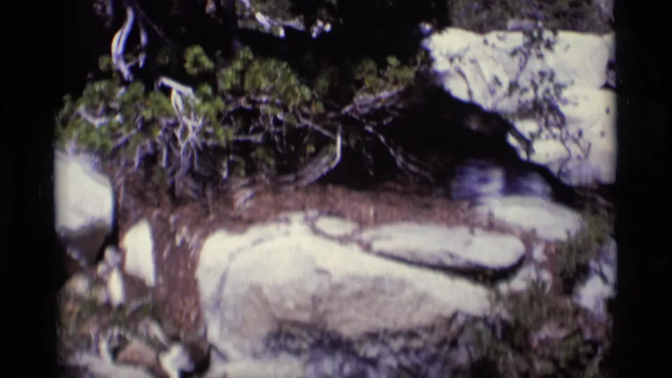 1975 8mm wilderness detail showing granite-like boulders, low vegetation, shaded soil, and a narrow stream channel.