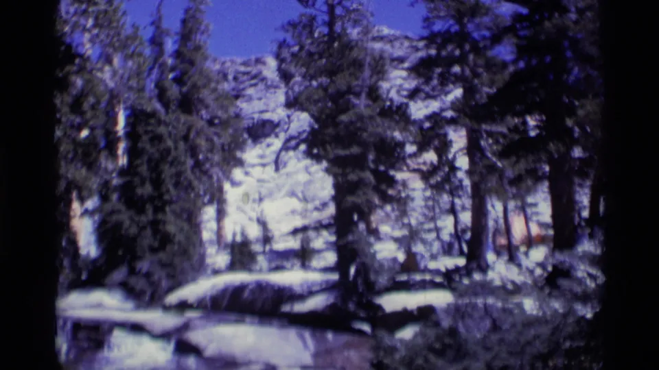1975 8mm view of a snowy evergreen forest in Minaret Wilderness, with rocky mountain ground and a clear blue sky.