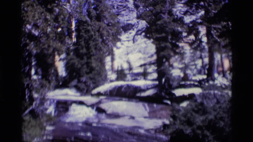 Minaret Wilderness forest scene from 1975 8mm film, showing snow-covered rocks, evergreens, and distant rocky terrain.