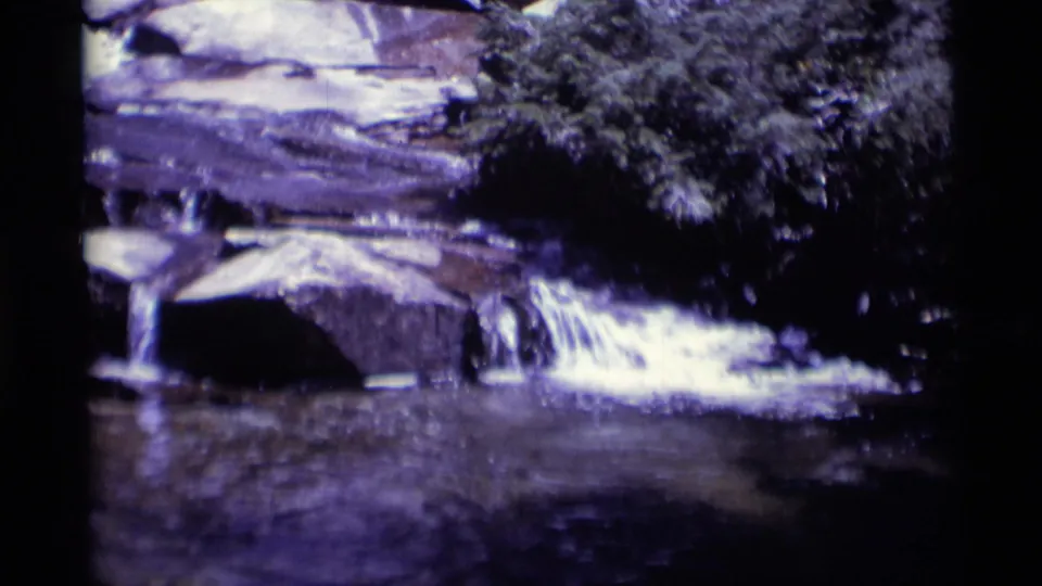 Aged 8mm frame showing water spilling over rocks near dense foliage and a dark forest pool in California wilderness.
