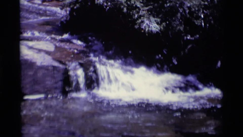 Vintage 8mm frame of a low California wilderness waterfall crossing flat rock with dense foliage overhead.