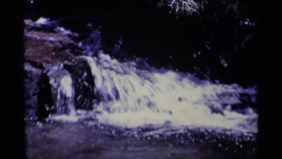 1970s 8mm wilderness view with foaming water rushing over dark rock ledges and a shaded stream below.