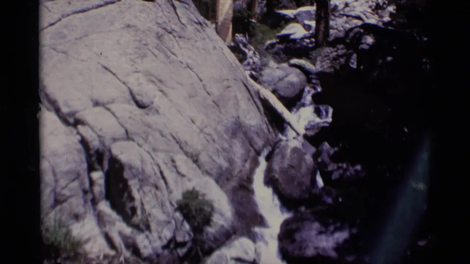Minaret Wilderness stream frame with rushing water pressed between pale granite and darker boulders in a handheld 8mm view.