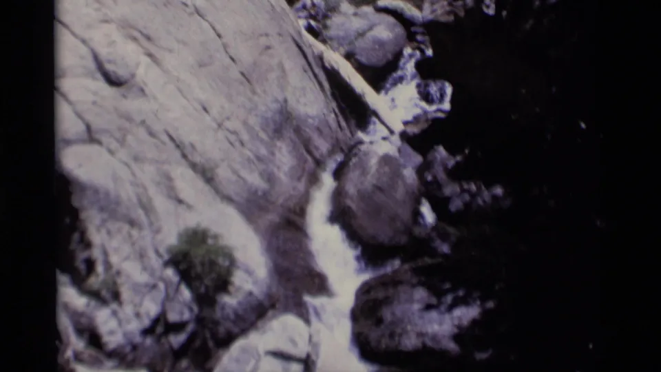 1975 wilderness home-movie frame showing a close rocky drop with bright water, pale stone, and sparse greenery near the lower edge.