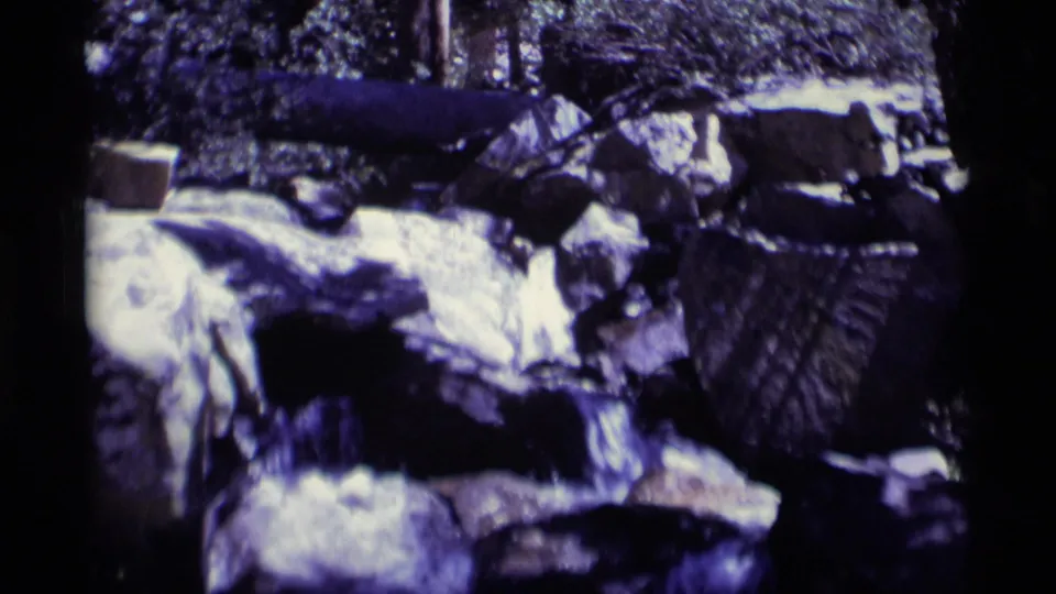 A grainy 8mm view of creek water crossing boulders in Minaret Wilderness, California, with trees and shaded brush behind.