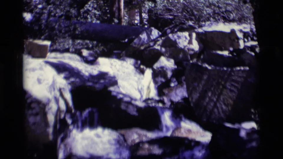 1975 home-movie footage frames a rocky creek bed with bright water, dark stones, and forest shade in Minaret Wilderness.