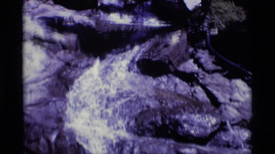 Aged 8mm footage shows foamy water flowing across flat rocks in a forested Minaret Wilderness stream.