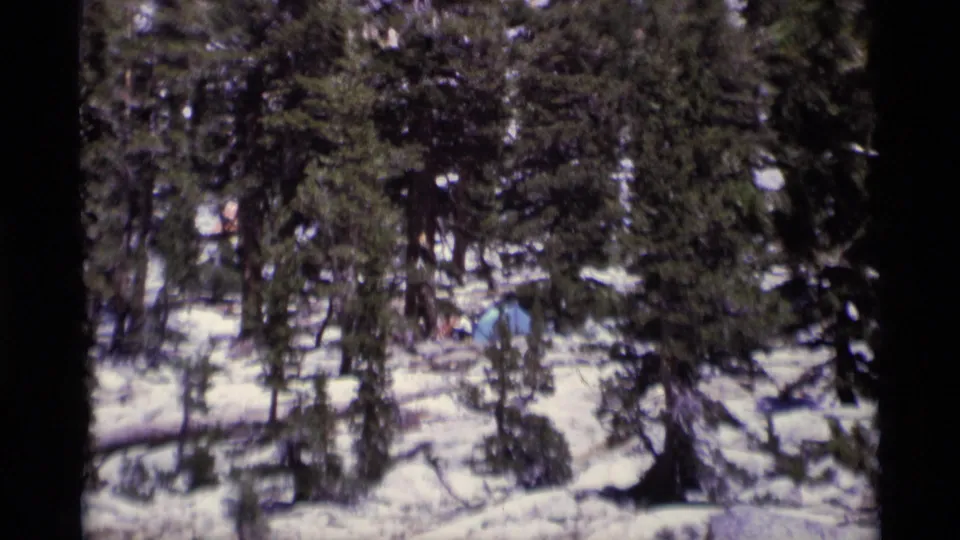 1975 Minaret Wilderness home-movie frame showing a blue tent and tiny distant campers among snowy conifers.