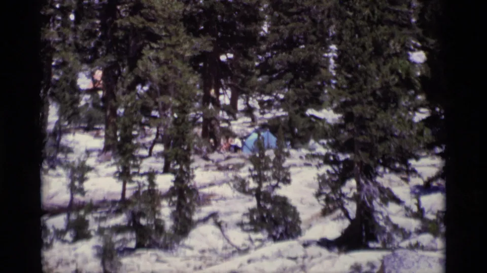 Zoomed 8mm frame of a snowy Minaret Wilderness campsite, with a blue tent and small people visible between conifers.