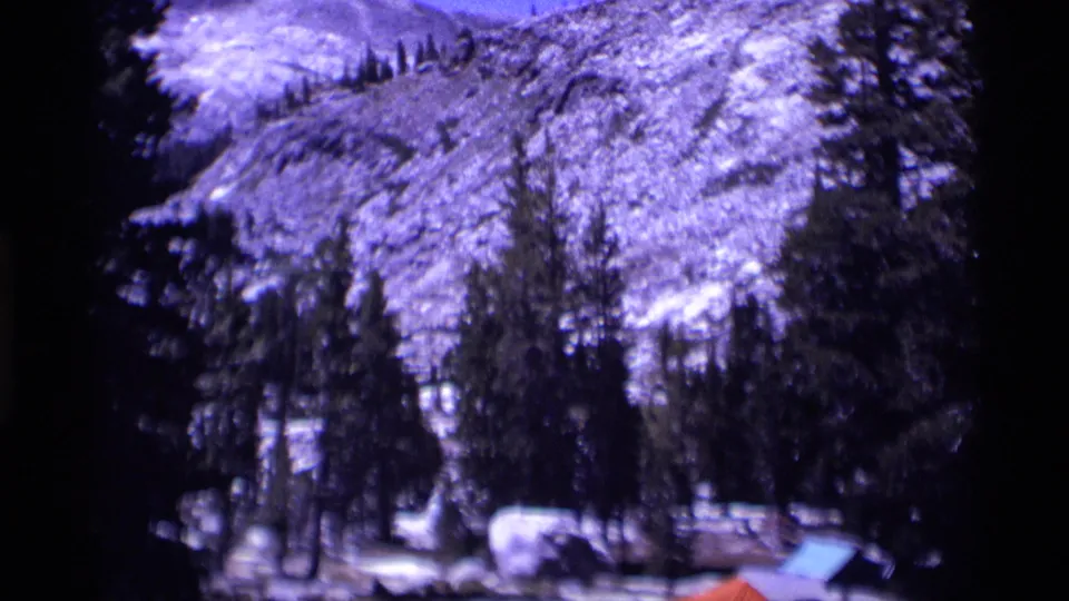 1975 Minaret Wilderness campsite framed by pines, with snow on the slope, a large boulder, and a blue tent among the trees.