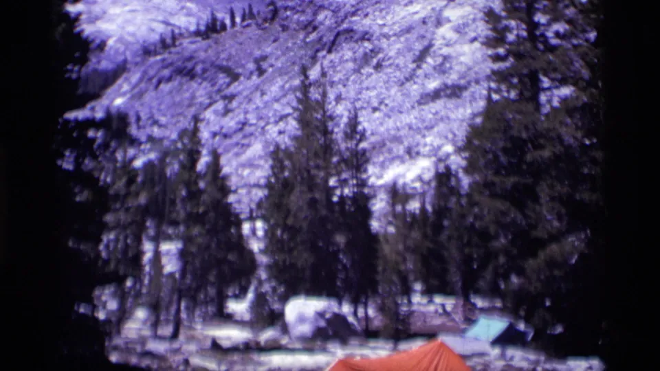 1975 8mm view of a Minaret Wilderness campground with pines, a large boulder, blue and orange tents, and snow-dusted terrain behind.