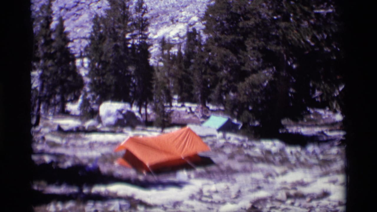 A remote Minaret Wilderness campsite shows an orange tent, green tarp, boulders, snow patches, and dense pines.