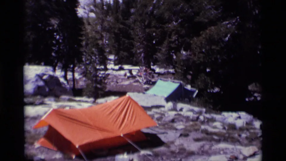 Closer 8mm view of an orange campsite tent and green tarp shelter in snowy, rocky Minaret Wilderness terrain.