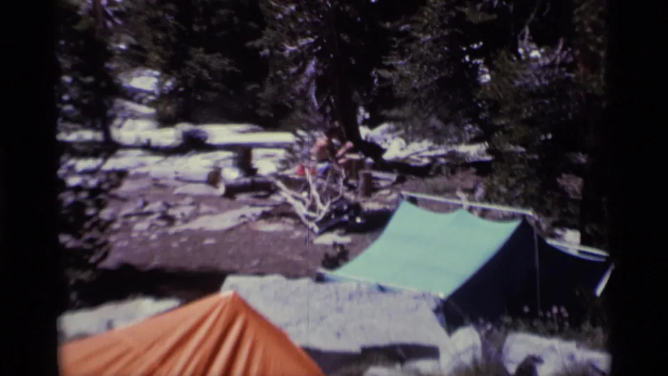 Campsite view with a green tarp shelter, edge of an orange tent, fallen branches, rocks, snow patches, and dark pines.