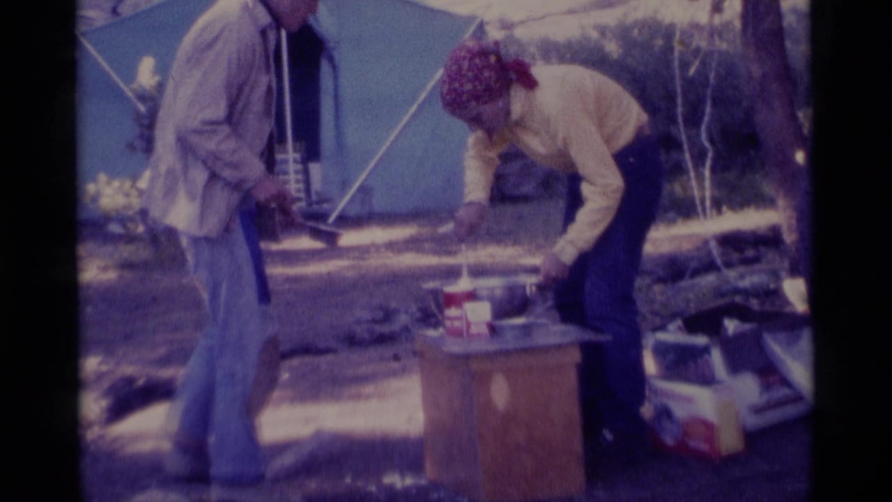 1975 Minaret Wilderness home-movie frame showing two adults near a blue tent as one bends over camp cookware on a small table.