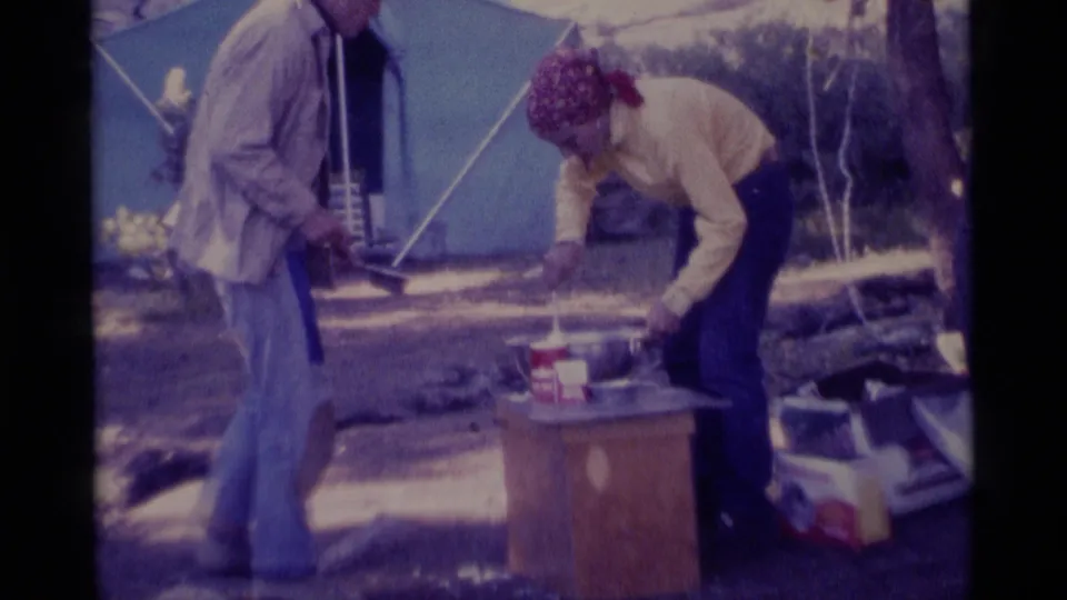 1975 Minaret Wilderness home-movie frame showing two adults near a blue tent as one bends over camp cookware on a small table.