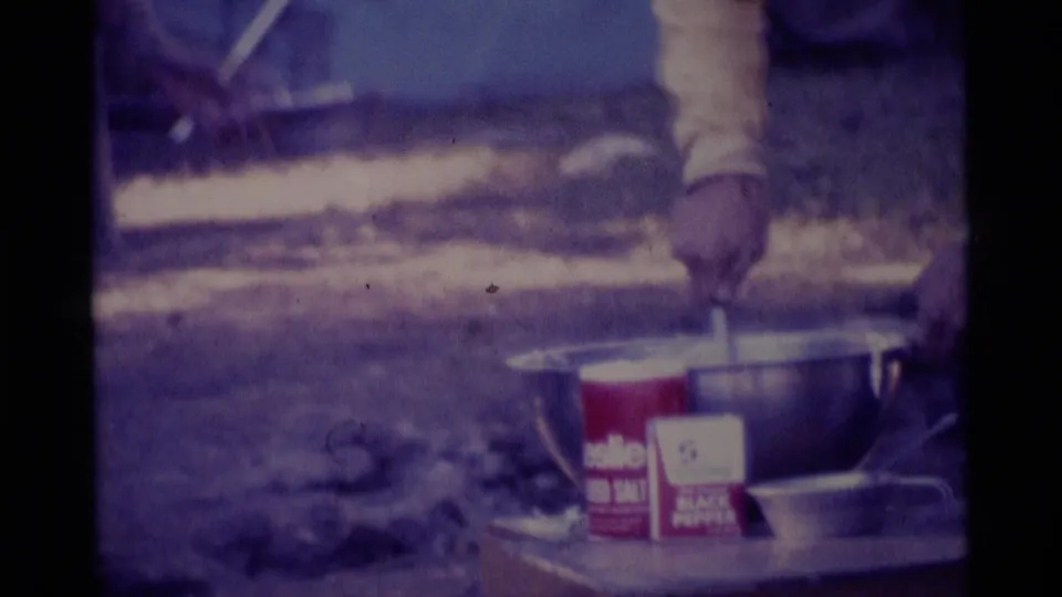 Close campsite frame showing a spoon stirring in a metal bowl with a red McCormick seasoning container visible on the makeshift table.
