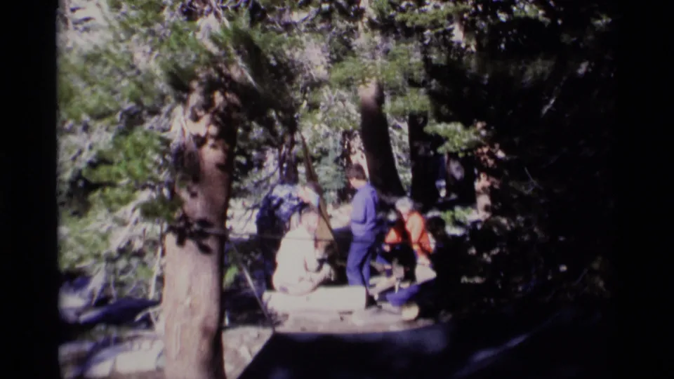 Wide 1975 home-movie frame showing a small group gathered among trees at a Minaret Wilderness campsite, with camping gear partly visible.