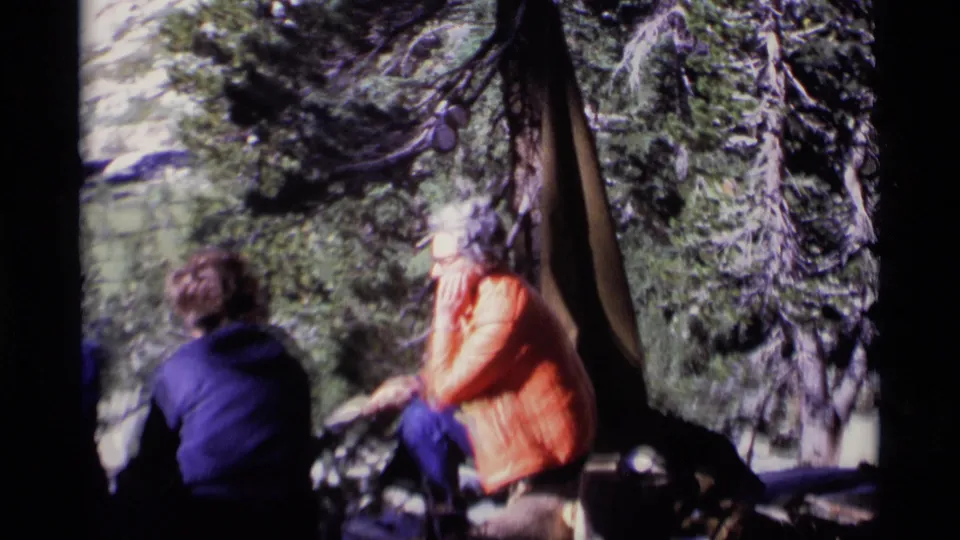 1975 8mm Minaret Wilderness frame showing campers seated by a tent among trees, with a woman in an orange jacket eating or resting.