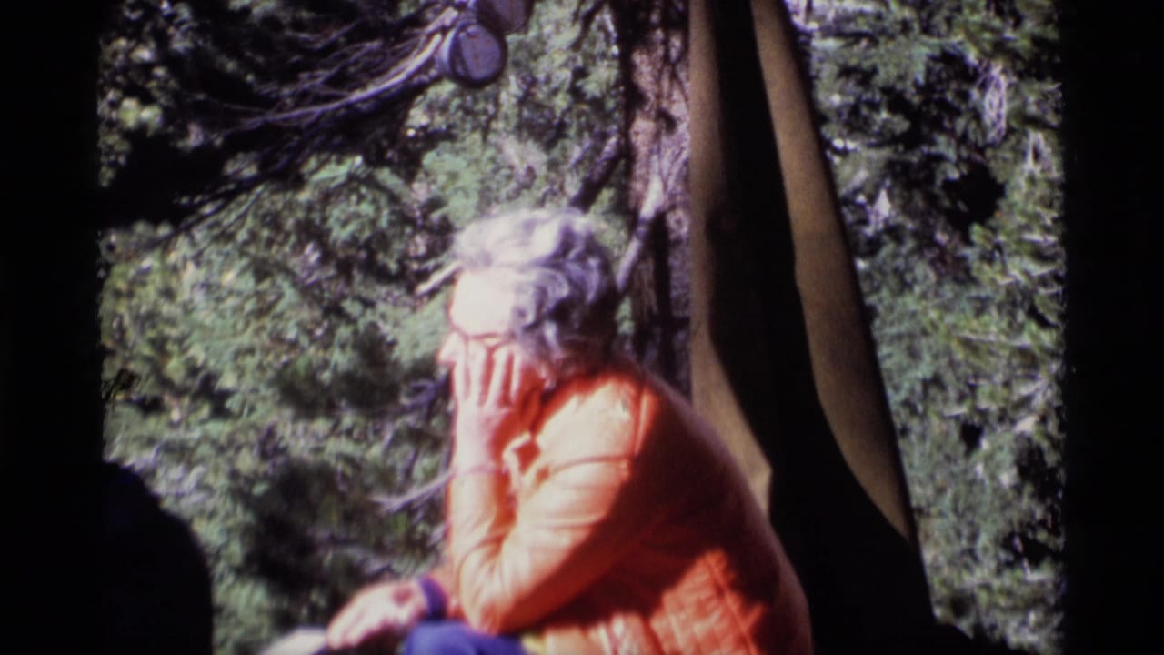 Woman in an orange jacket sits beside tent fabric in a wooded campsite.