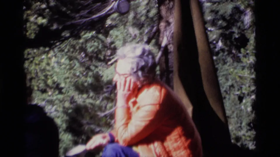 8mm frame from the 1975 Minaret Wilderness camping scene showing a woman seated near tent fabric, surrounded by forest shade.