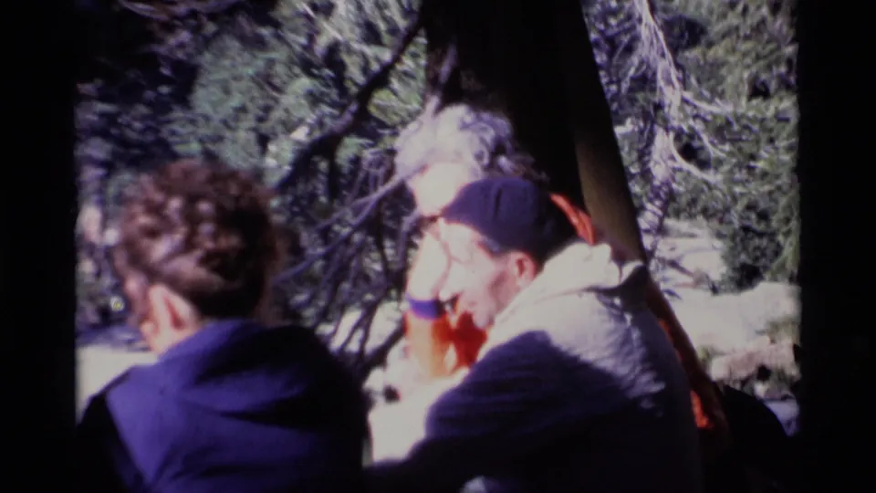 Close 1975 forest frame with seated companions and an older man in a cap near bright pine branches.