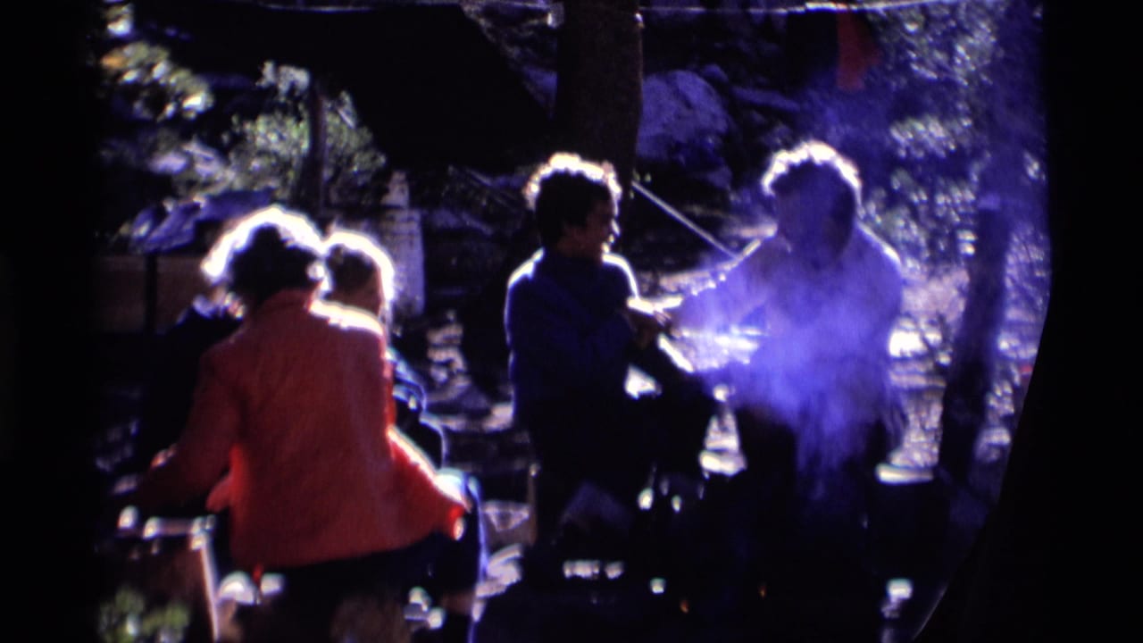 1975 8mm frame from Minaret Wilderness showing adults seated around a smoky campfire among trees and rocks.